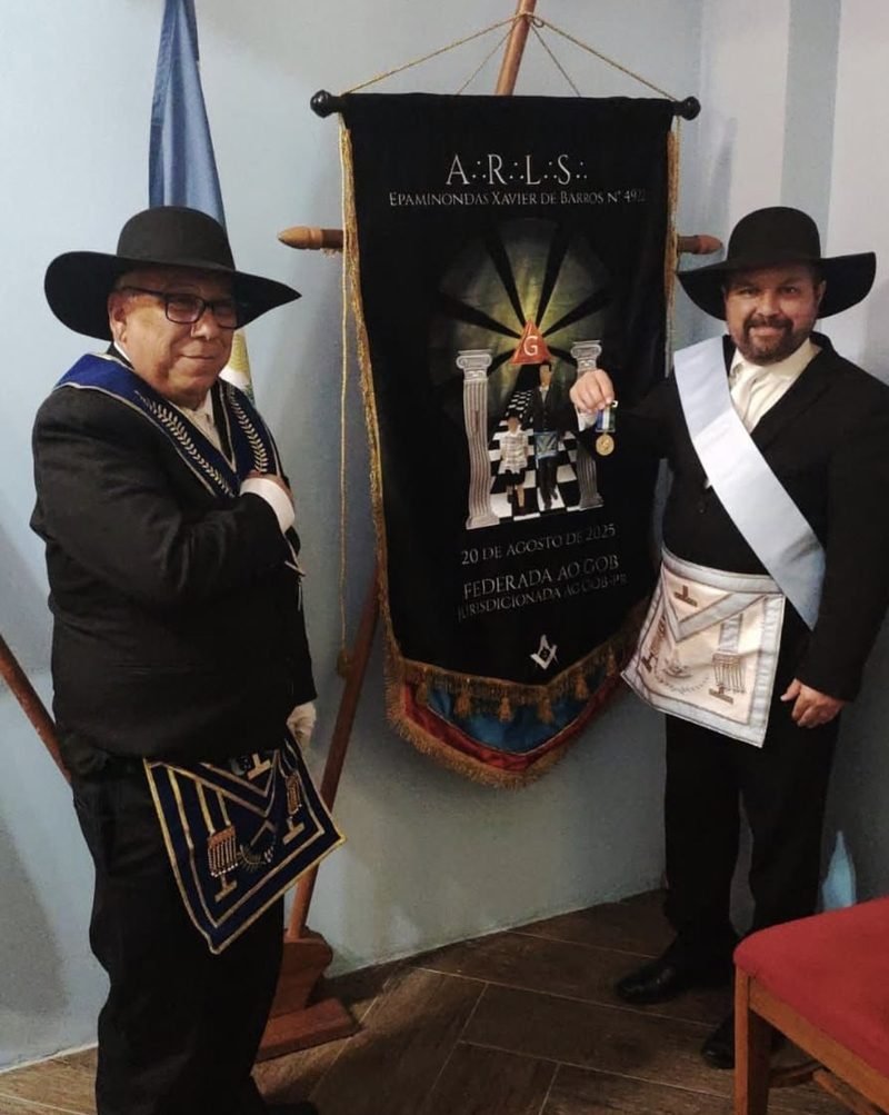 Dois irmãos maçons, com chapéu e avental, ao lado do estandarte da A∴R∴L∴S∴ Epaminondas Xavier de Barros