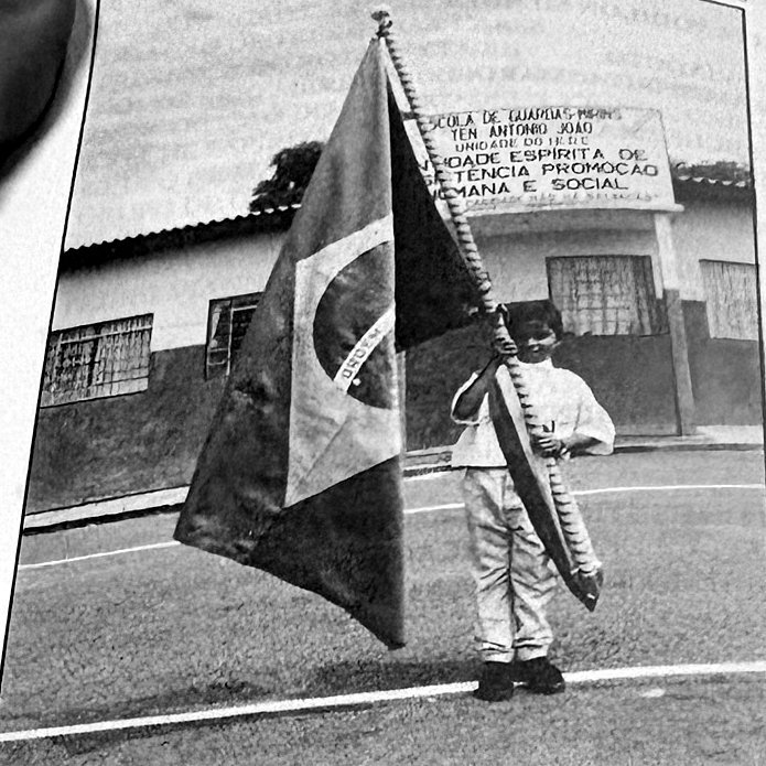 Menino segura a Bandeira Nacional em frente à fachada da Escola de Guardas Mirins Ten. Antônio João